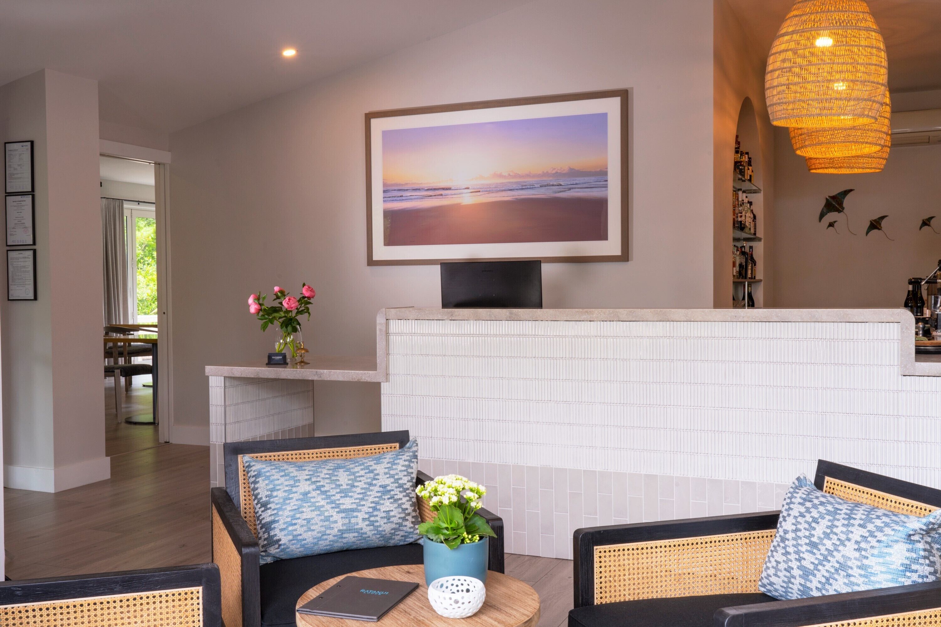 A modern living room with a large flat-screen TV, wicker furniture, and a view of the beach through the window.