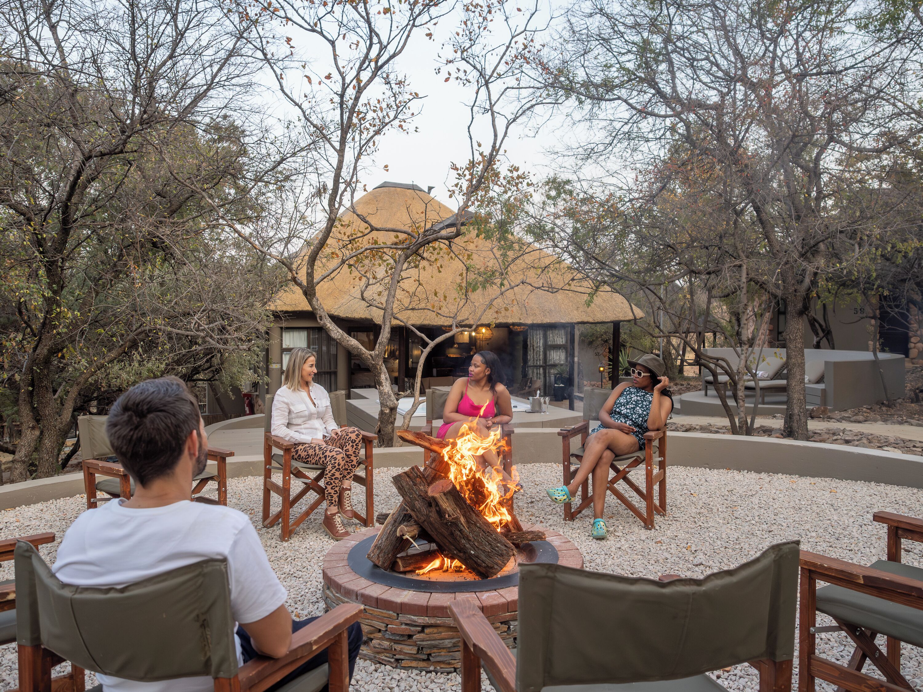 A group of people seated around a fire pit in an outdoor setting with trees and a building in the background.