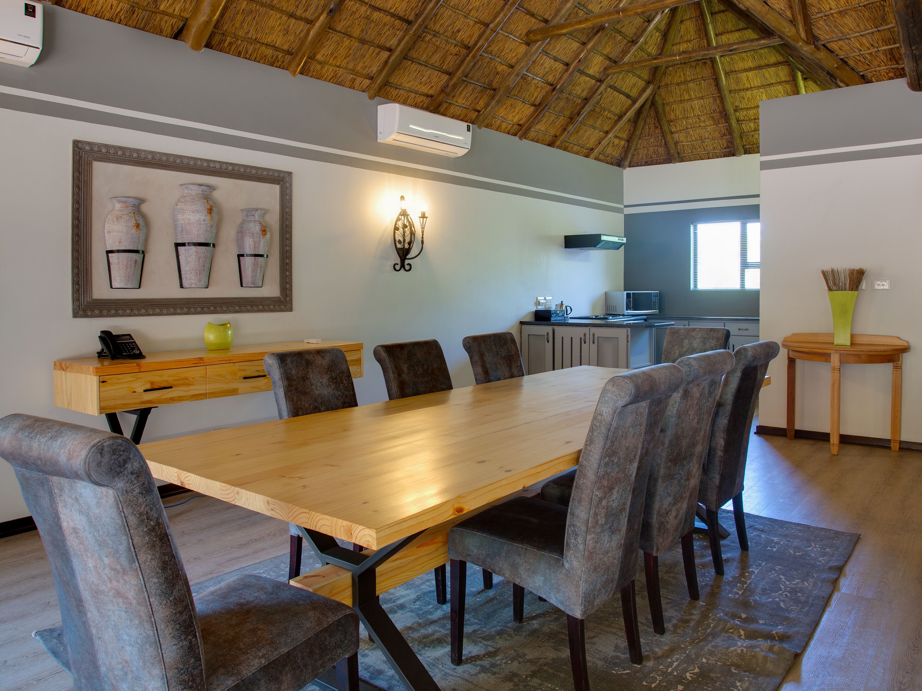 Uma sala de jantar com mesa e cadeiras de madeira, cozinha ao fundo e teto de colmo.