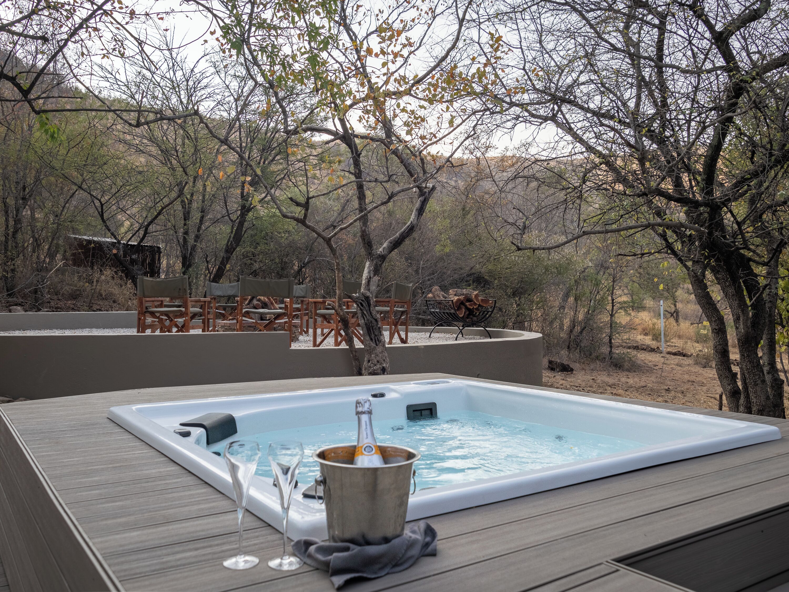A hot tub surrounded by wooden deck chairs and a fire pit, set in a natural outdoor area with trees and a hill in the background.