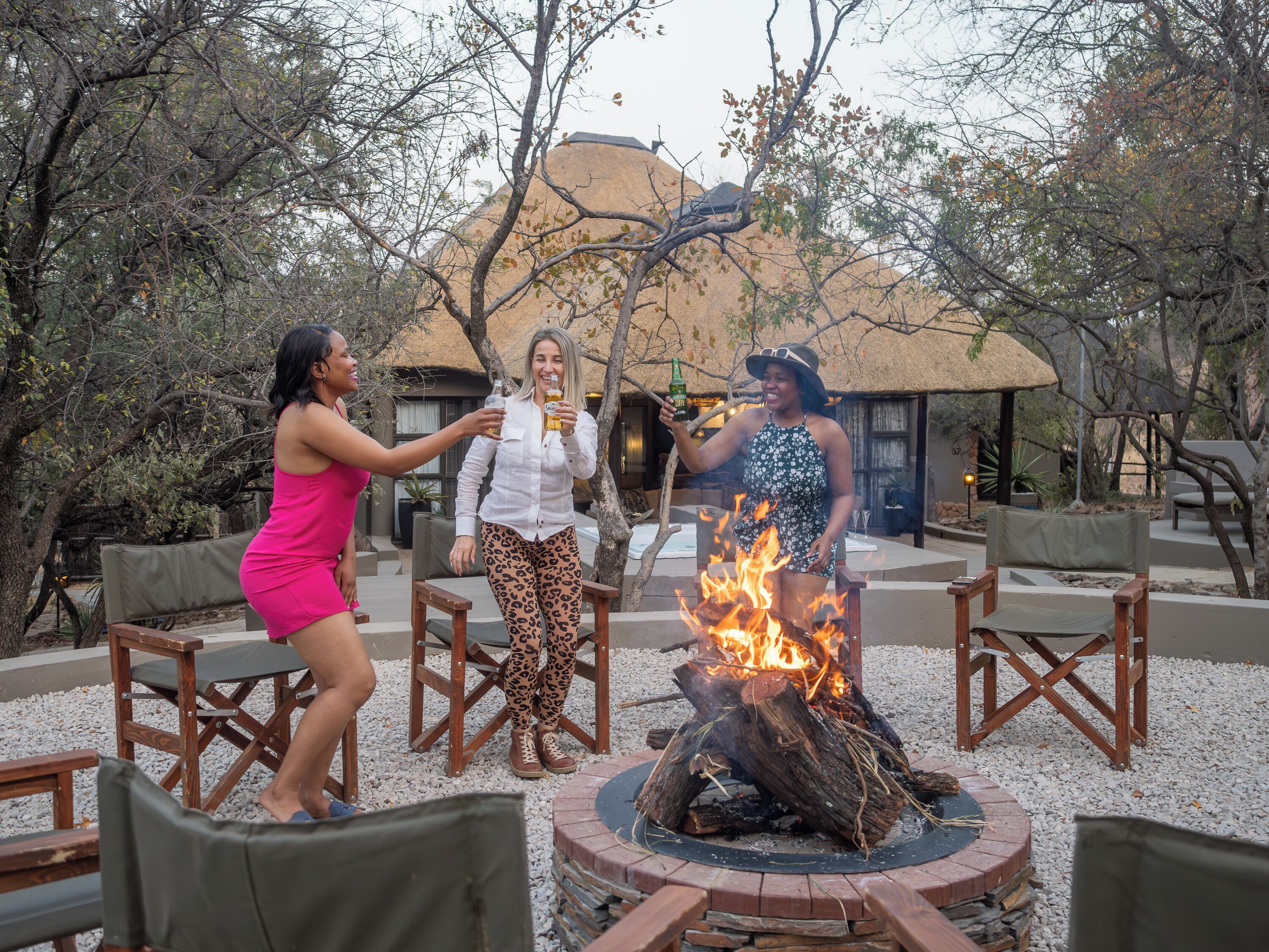 Three people around a fire pit with a thatched-roof building in the background.
