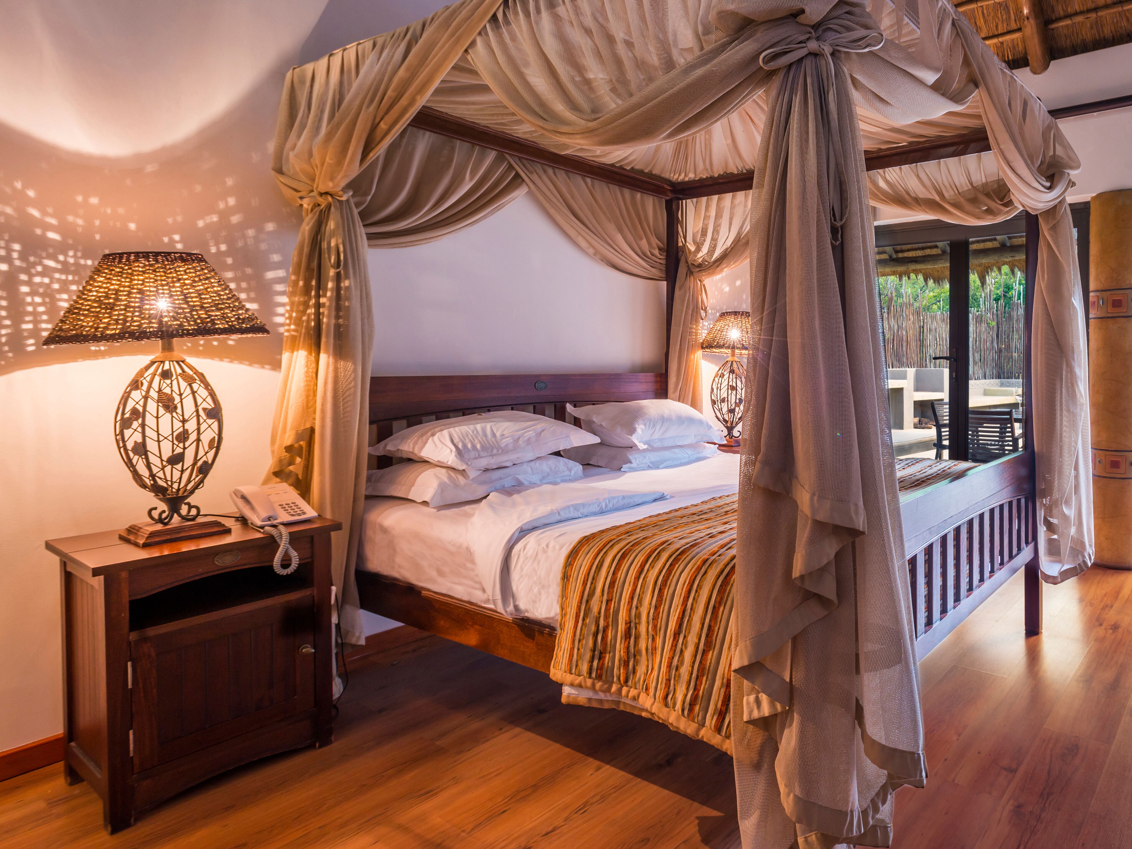 Un lit à baldaquin à quatre poteaux avec une tête de lit en bois, une table de chevet avec une lampe, et une vue sur un espace extérieur à travers le baldaquin.