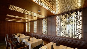A restaurant with wooden paneling, cushioned booths, and decorative screens. Tables are set with white tablecloths, plates, and cutlery. - Hotel Regent Grand (New Delhi)