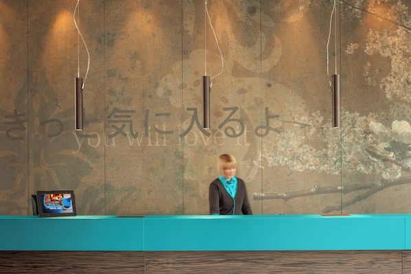 A reception desk with a digital screen displaying a hotel image, a wooden wall with Japanese characters, and a teal-colored counter. - Motel One Düsseldorf-Hauptbahnhof (Düsseldorf)