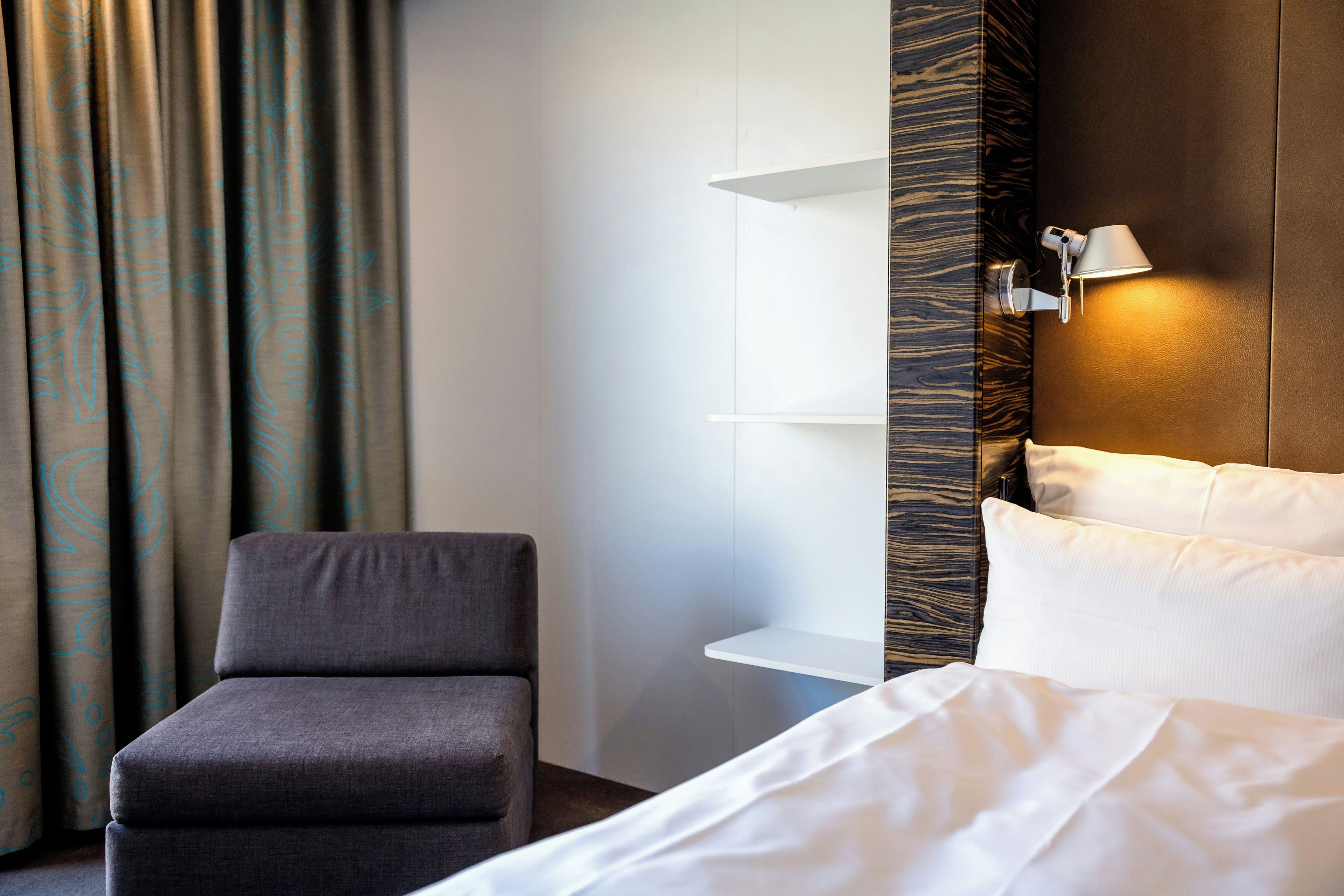 A hotel room with a bed, a chair, a nightstand, and a wall-mounted lamp.