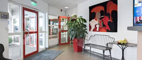 A hotel lobby with a reception desk, a bench, a large potted plant, and a wall-mounted screen displaying information.