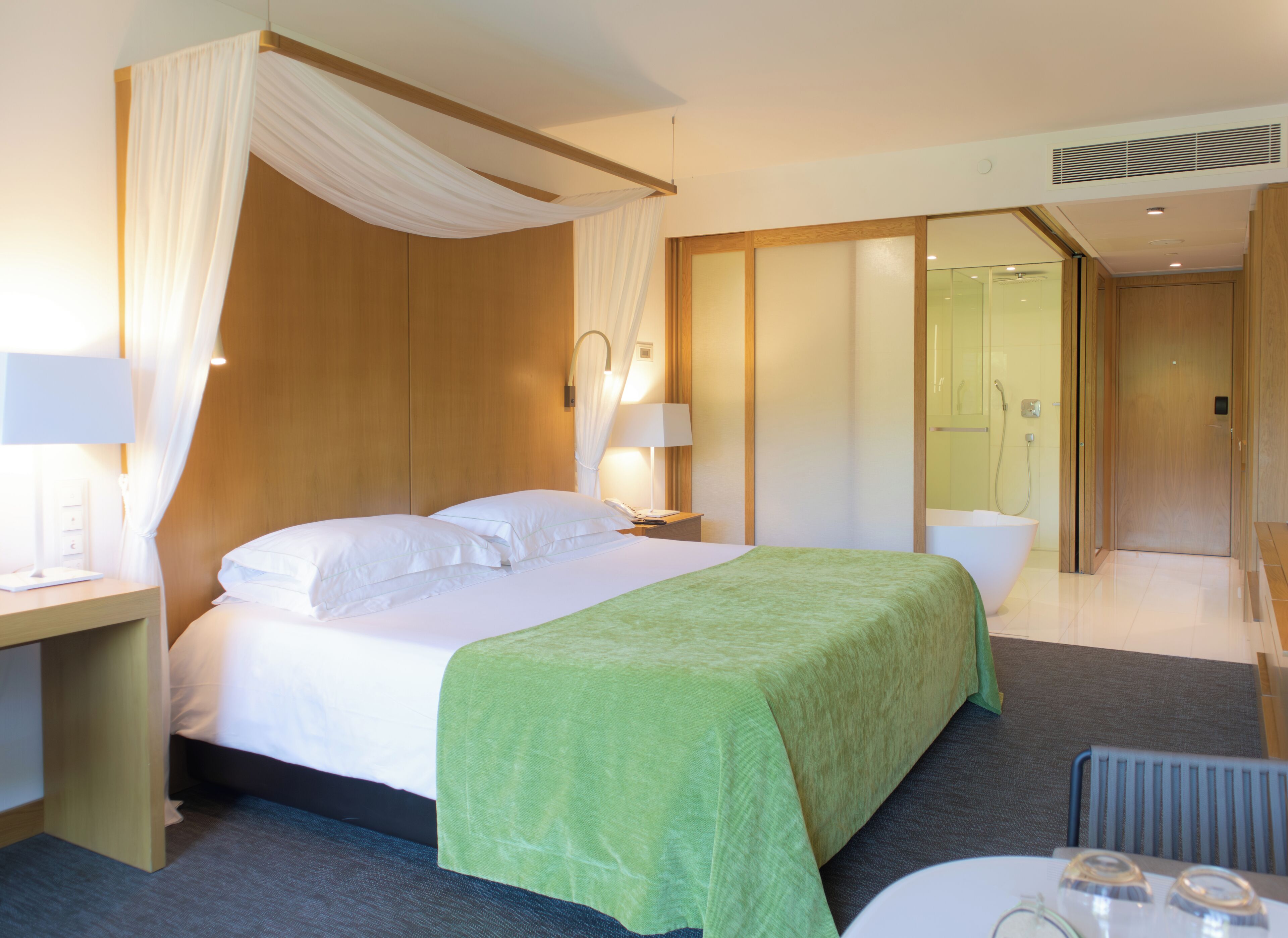 Ein Hotelzimmer mit einem Bett, einem Nachttisch, einer Lampe, einer grünen Tagesdecke, einer Duschkabine und einem Glaskastenbad.