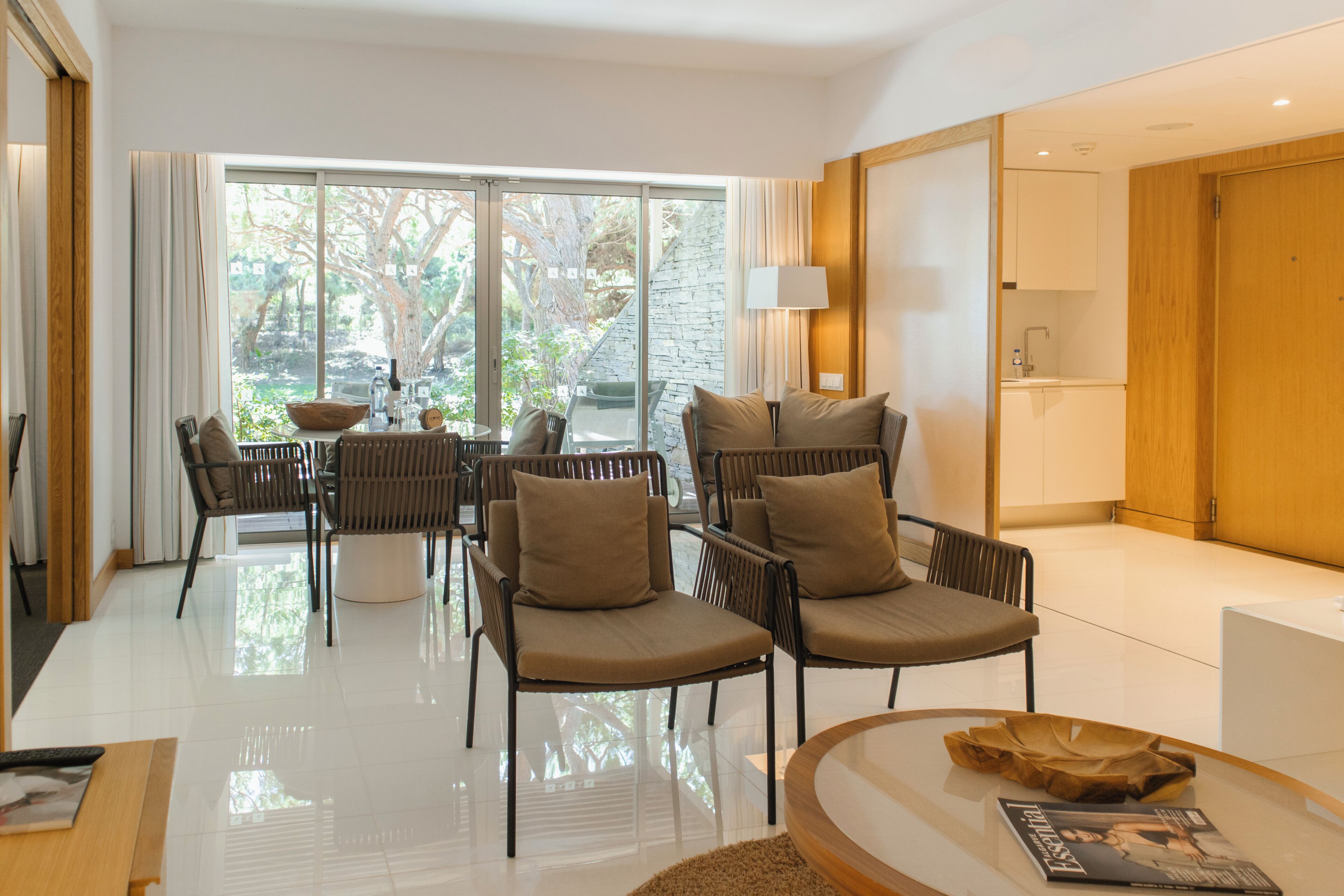 A modern living room with a glass coffee table, comfortable seating, a view through large windows, and a wooden door.