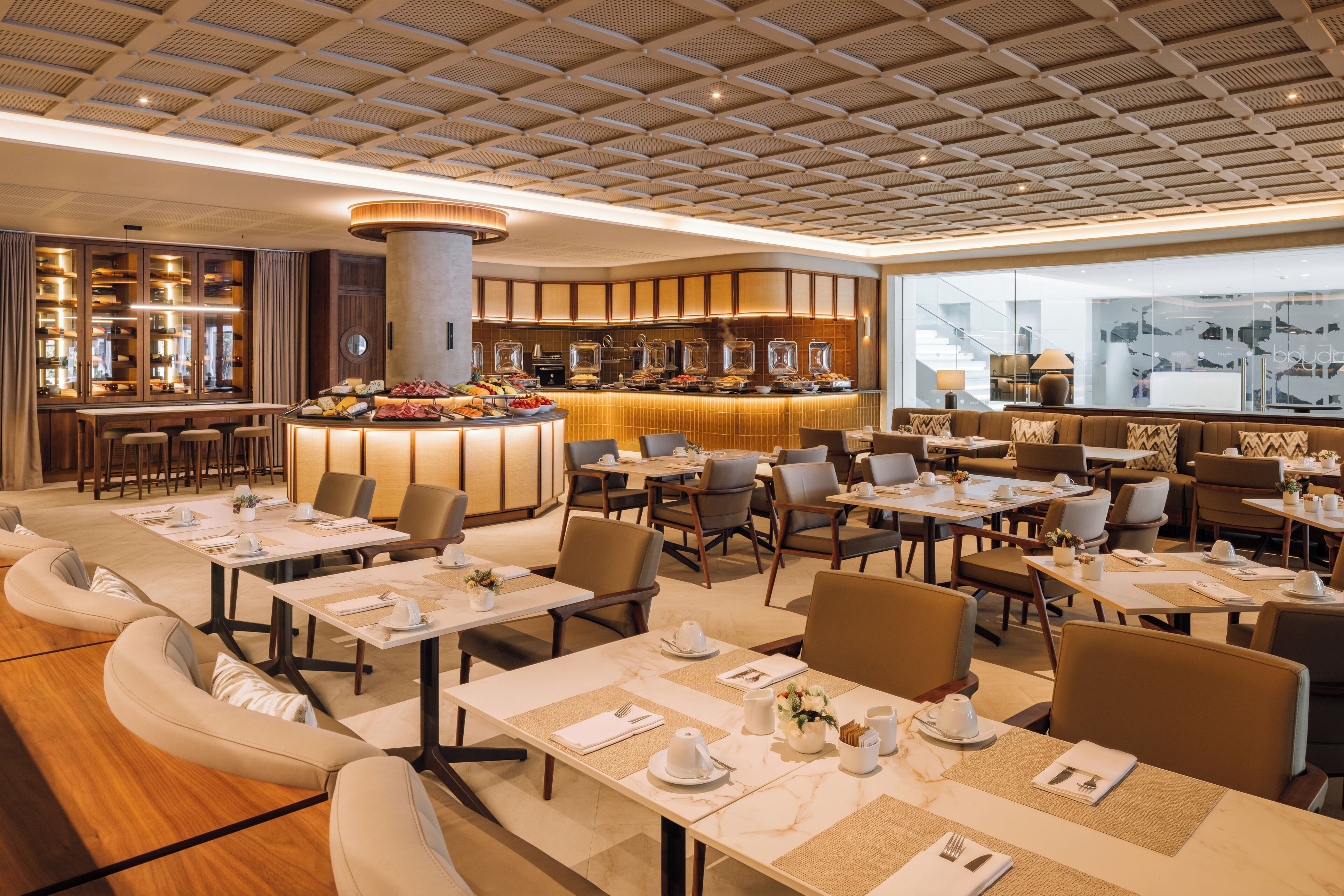 A hotel dining area with a central buffet, multiple dining tables, and a bar.