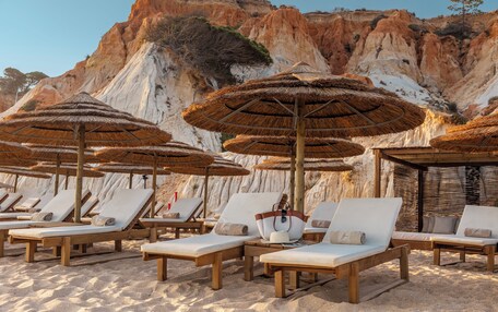 A beach with wooden lounge chairs and thatched umbrellas, set against a backdrop of rocky cliffs.