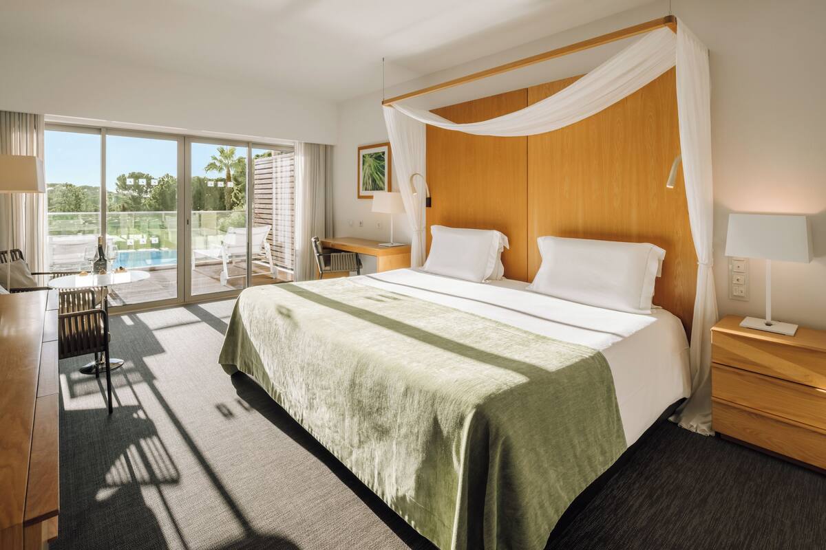 A hotel room with a large bed, a desk, a chair, a nightstand, and a view of an outdoor pool area.