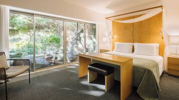 Una habitación de hotel moderna con un gran ventanal corredero, un escritorio de madera, una cama con ropa blanca y vistas a zonas verdes.