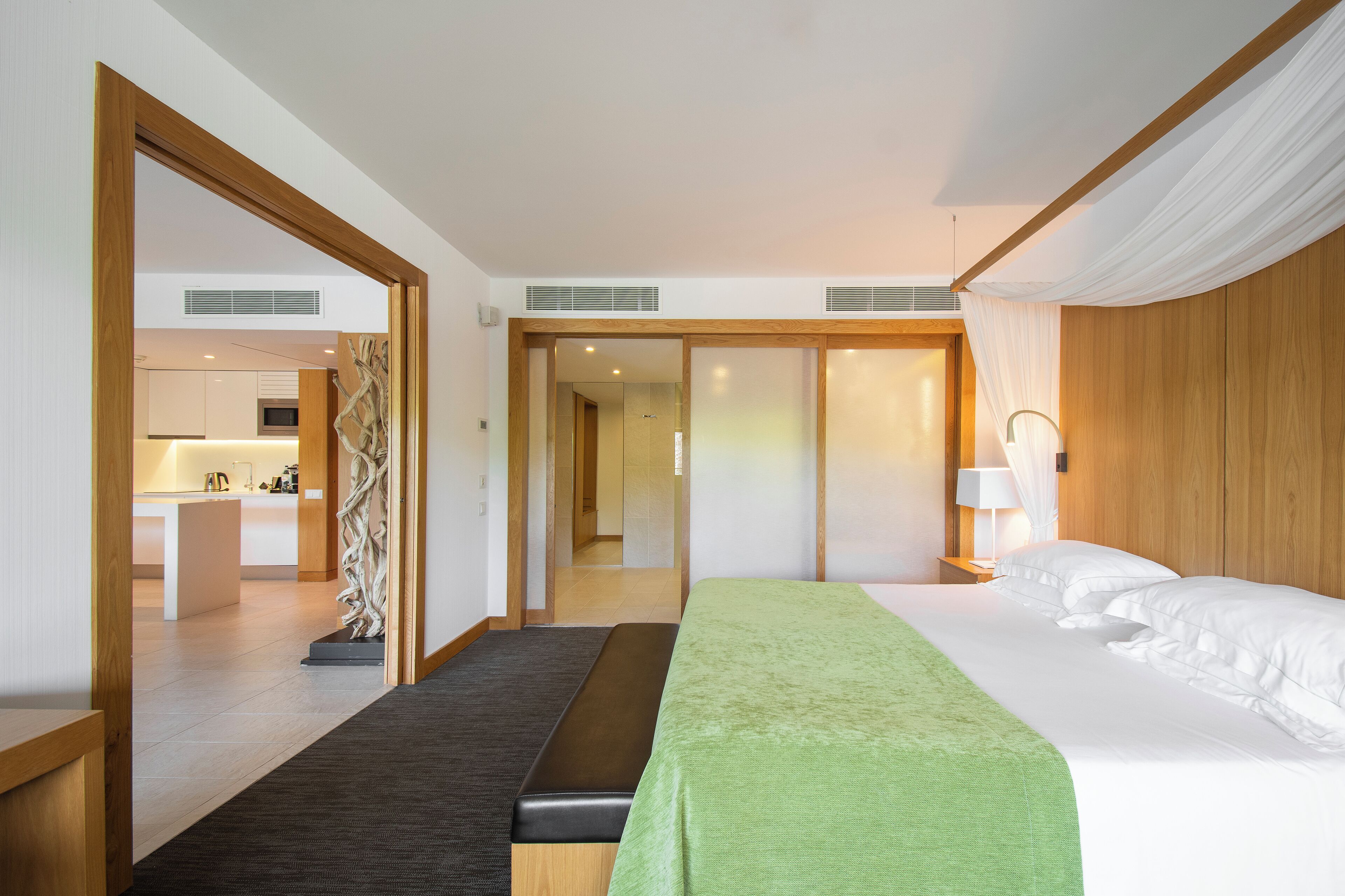 A hotel room with a bed, bedside table, lamp, and a view of the kitchen.
