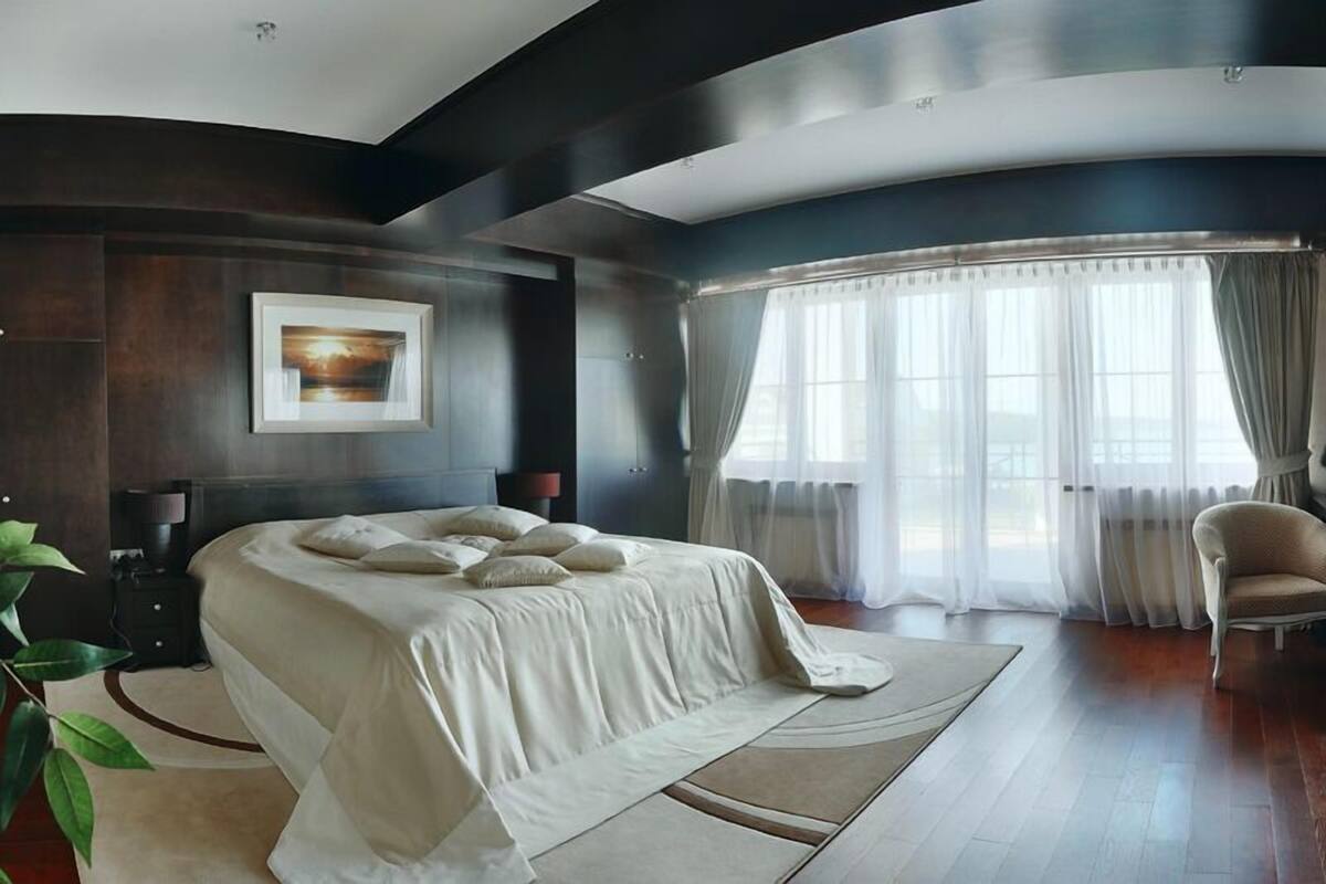 A modern bedroom with a large bed, a nightstand, a lamp, a chair, and a large window with curtains.