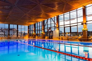 Une piscine intérieure à l’eau claire, entourée de grandes fenêtres et ornée de poutres en bois au plafond.