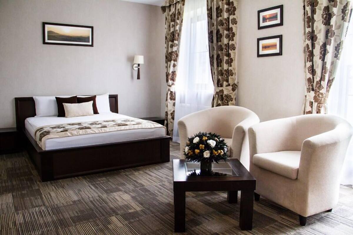 A hotel room with a bed, two armchairs, a small table with a flower vase, and a wall-mounted lamp.