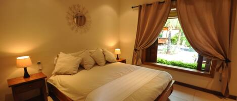 A bedroom with a large bed, two bedside tables with lamps, a window with curtains, and a wall-mounted mirror.