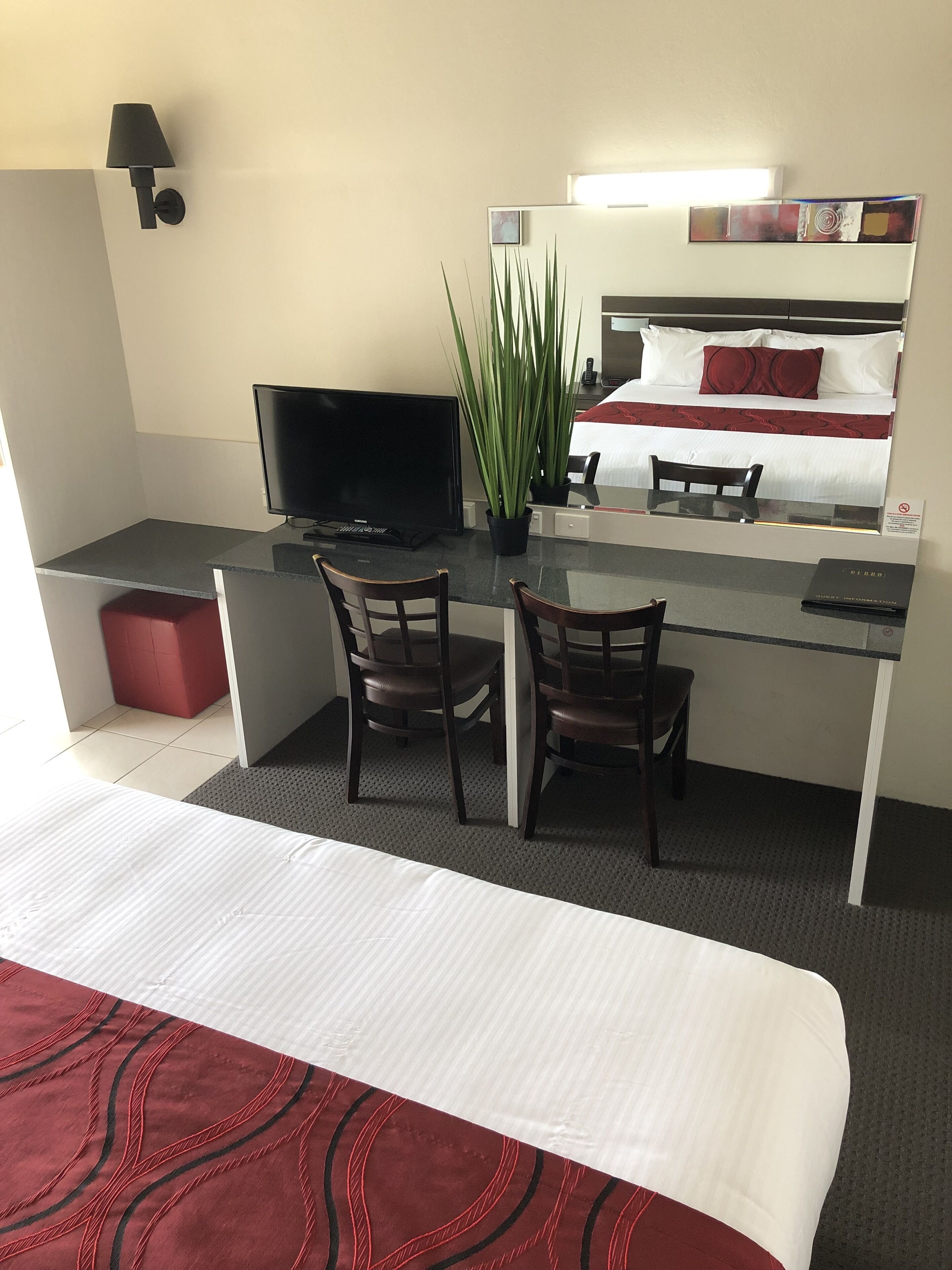 A hotel room with a bed, a desk with two chairs, a television, a potted plant, and a mirror reflecting the bed and a red pillow.
