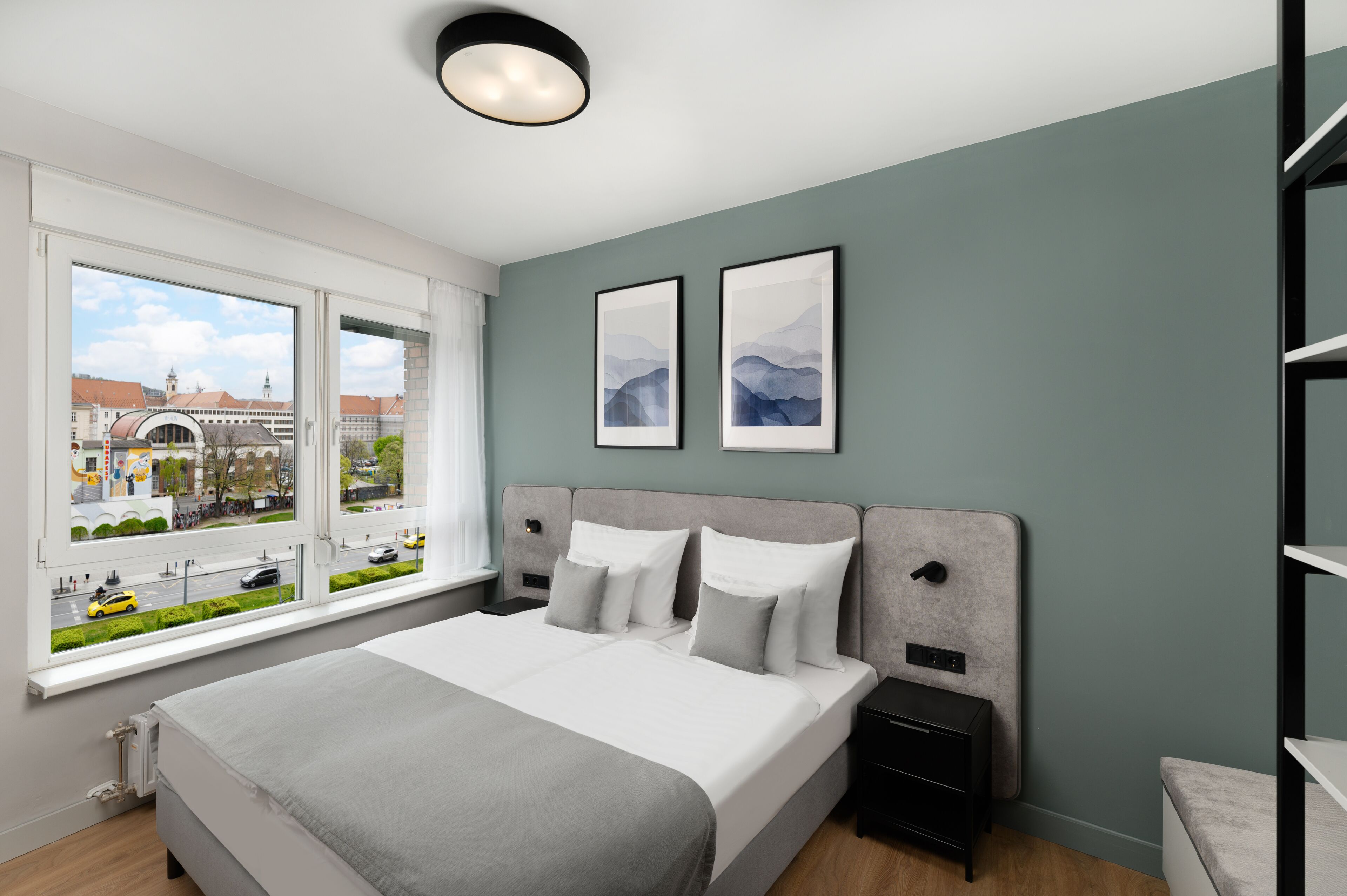 A modern hotel room with a large bed, two bedside tables, a nightstand, and a window offering a city view.