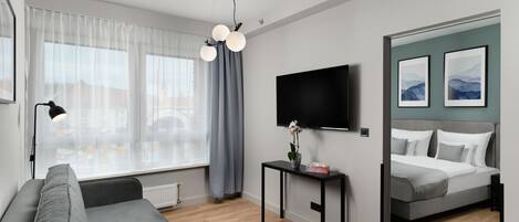 Una habitación de hotel moderna con un sofá gris, una pequeña mesa con un jarrón y una caja, una cama con ropa de cama gris, un televisor montado y un ventanal con cortinas translúcidas.