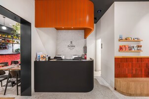 Ένας πάγκος υποδοχής με μοντέρνο σχεδιασμό, ένας γκρι τοίχος με την επωνυμία του ξενοδοχείου και ένας τοίχος επενδυμένος με κόκκινα πλακάκια.