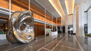 Een moderne lobby met een groot reflecterend kunstwerk, gepolijste marmeren vloeren en een ruim interieur met een receptiebalie en zitgedeeltes.