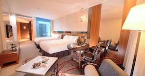 A hotel room with two beds, a sitting area with a sofa and armchairs, a small table with a laptop and a cup, and a TV on the left.