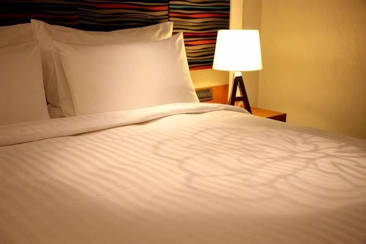 Cama bem arrumada com lençóis e almofadas brancas, uma luminária de cabeceira e um padrão de parede listrado ao fundo.