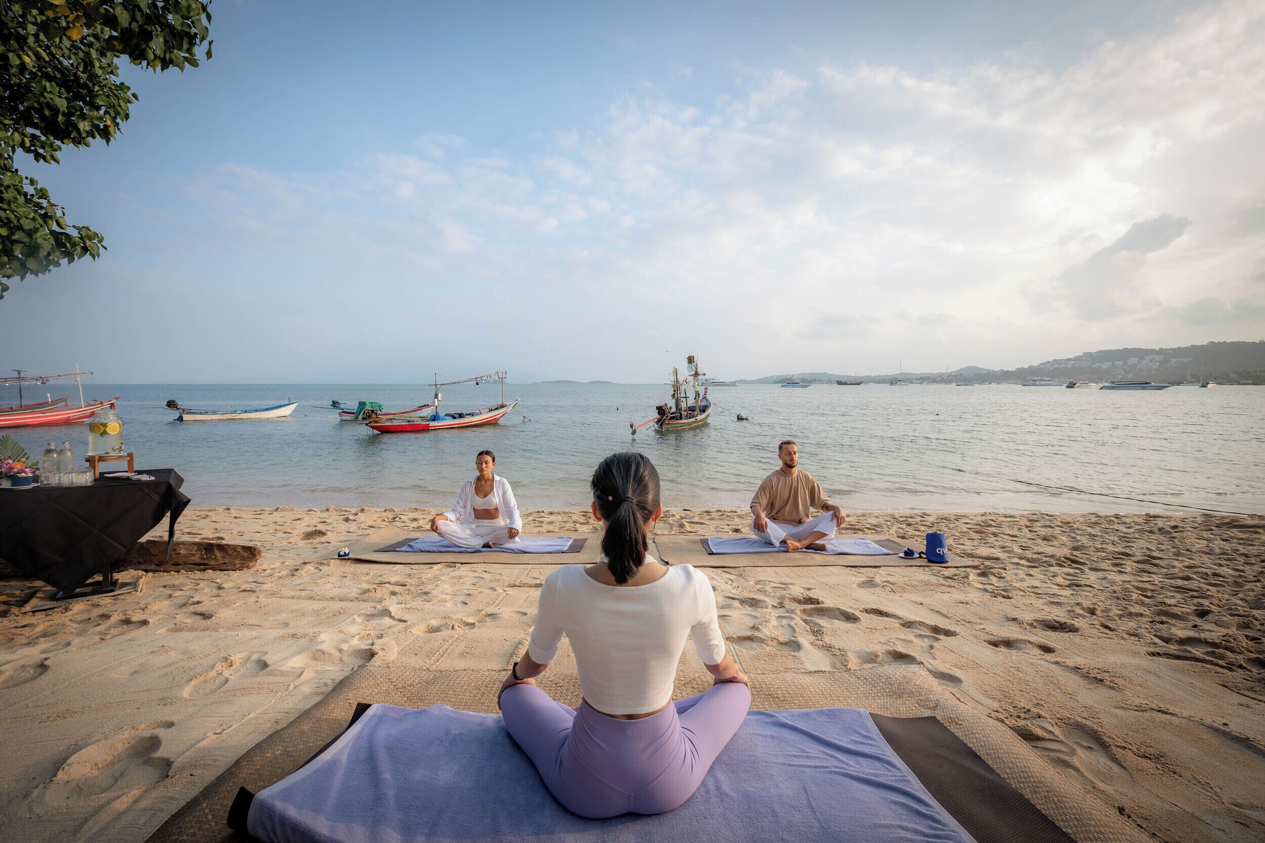 Trois personnes méditent sur des tapis posés sur une plage, avec des bateaux sur l’eau.