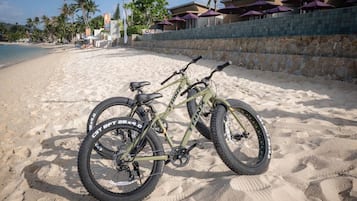 A beachside bicycle with a chain guard and a front suspension fork, parked on sandy beach with a stone wall and umbrellas in the background.