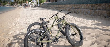 A beachside bicycle with a chain guard and a front suspension fork, parked on sandy beach with a stone wall and umbrellas in the background.