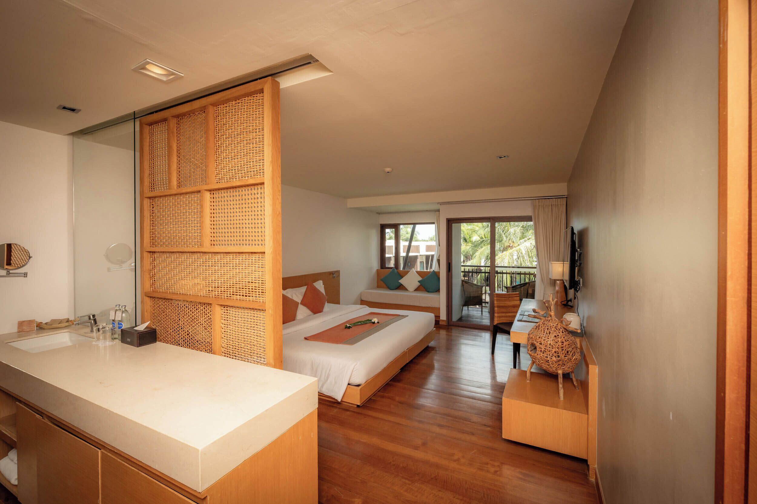 A modern hotel room with a large bed, a separate sitting area, a bathroom with a large mirror, and a wooden floor.