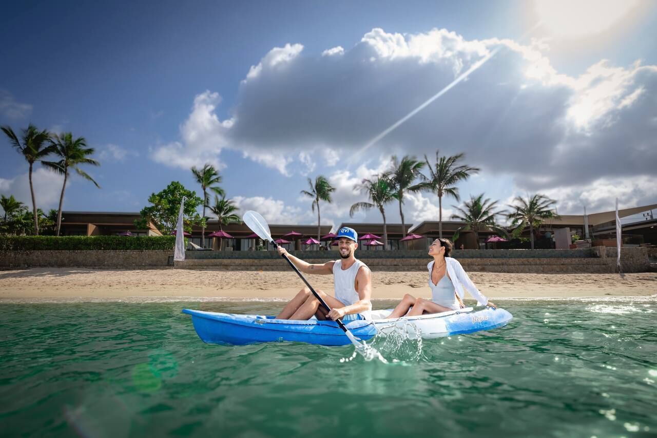 Un couple est assis dans un kayak bleu, et l’on aperçoit des palmiers et un complexe hôtelier en arrière-plan, par une journée ensoleillée.