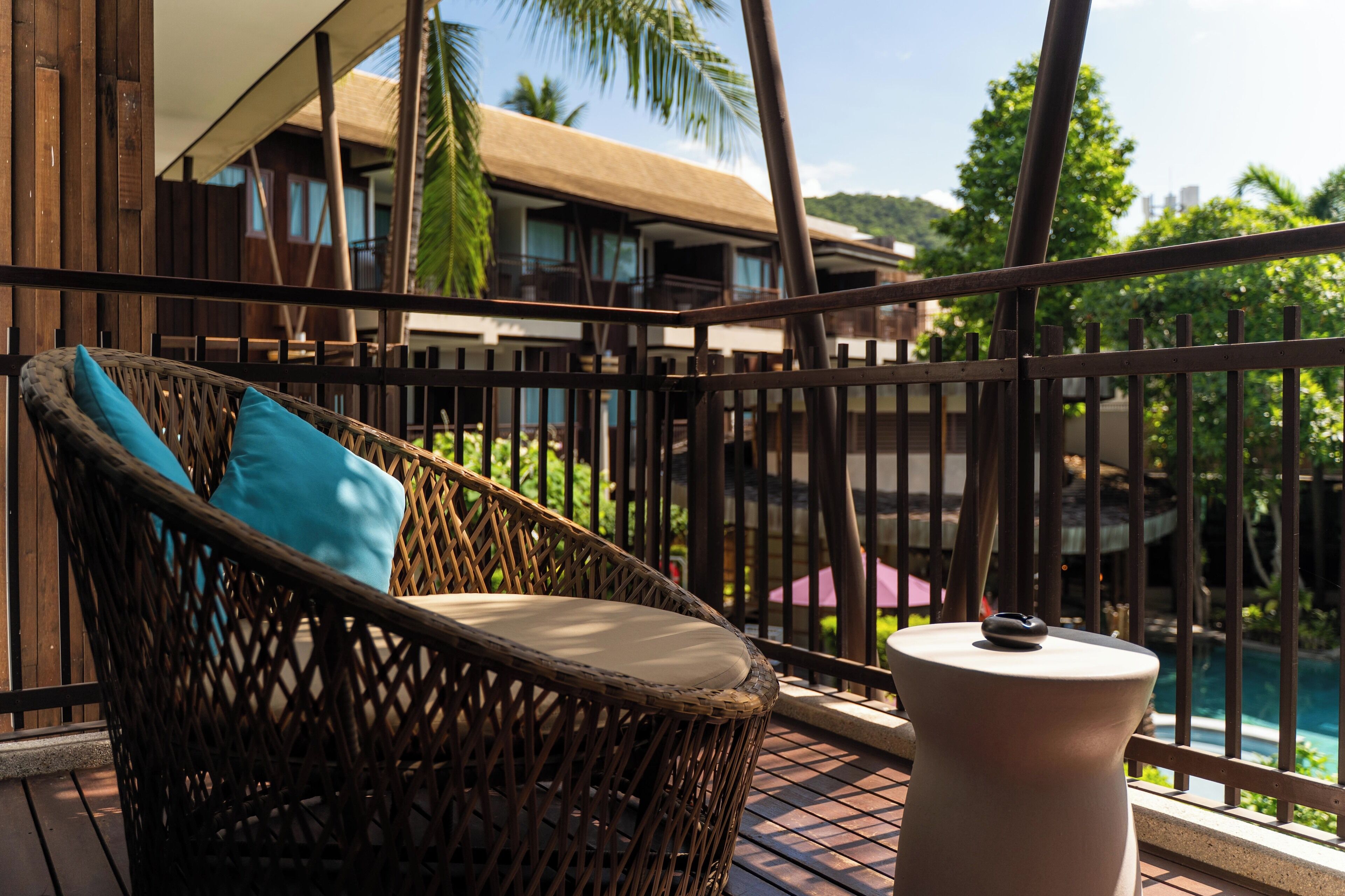 Une chaise en osier avec un coussin et une petite table sur un balcon donnant sur une piscine et des immeubles.