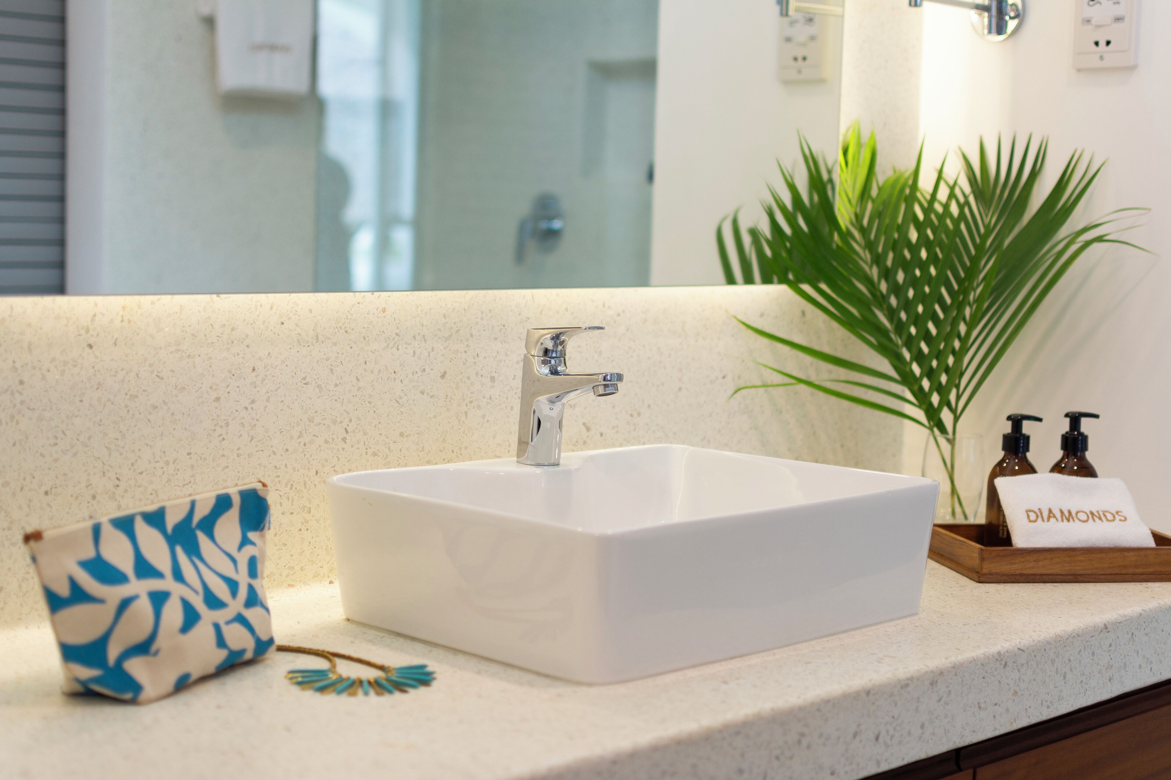 A bathroom with a white sink, a small blue and white patterned bag, a plant, and a tray with two bottles and a towel labeled "Diamonds".