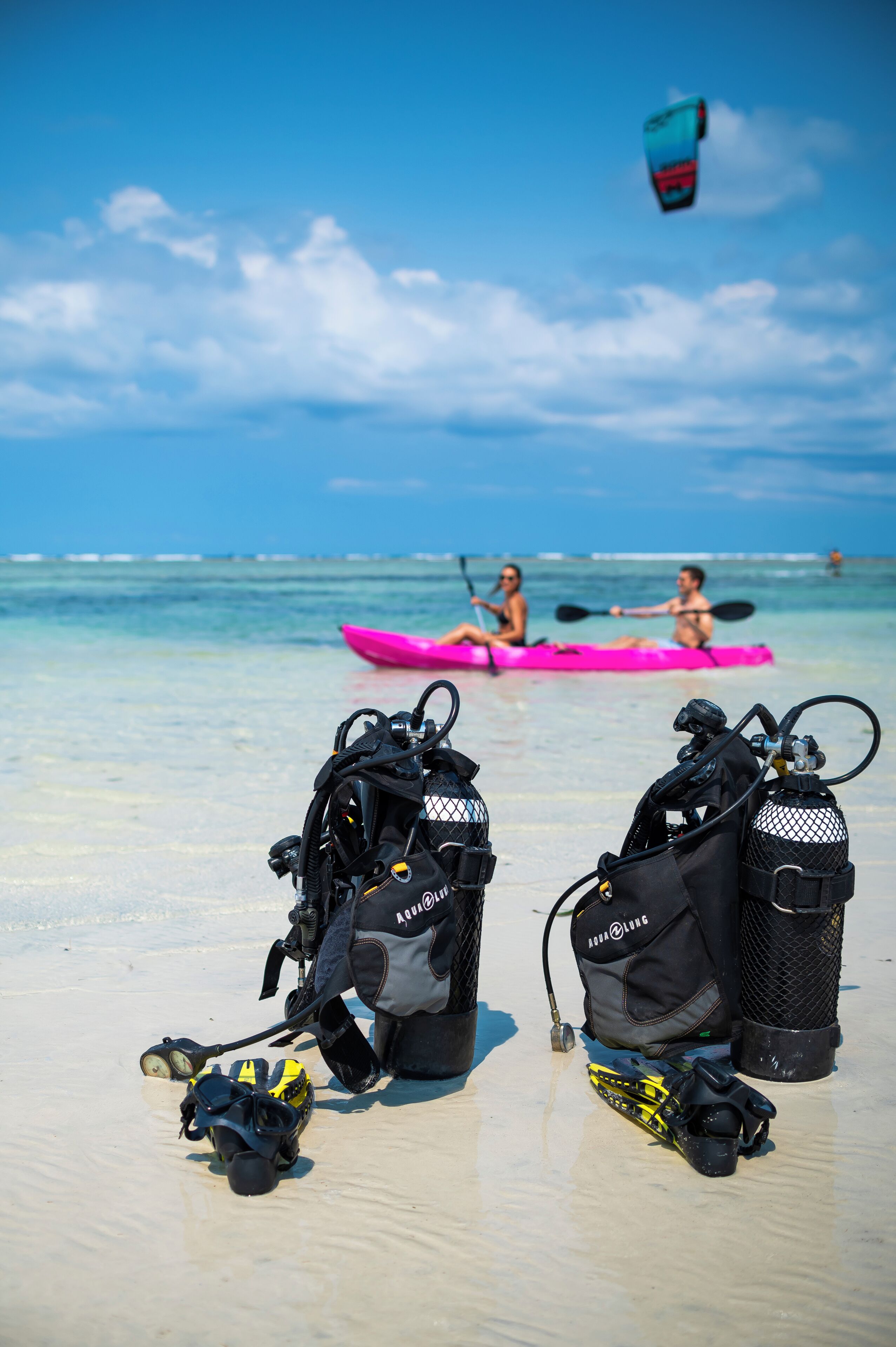 Equipo de buceo en una playa arenosa, con kayakistas al fondo.