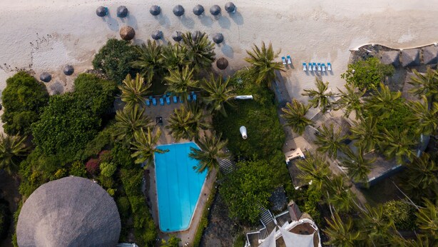 Vue aérienne d’un complexe hôtelier de bord de mer, avec une piscine, des chaises longues et des palmiers.