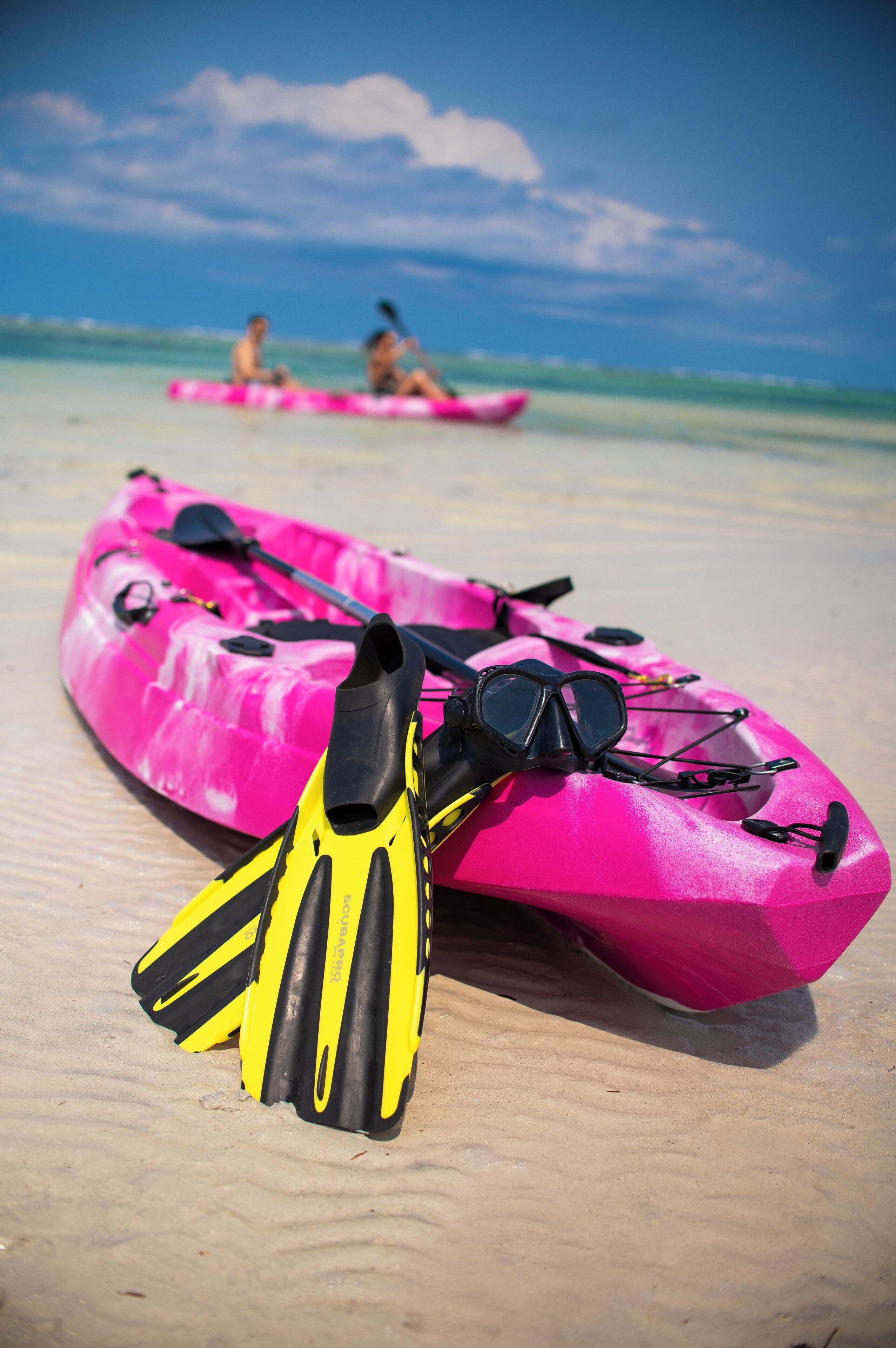 A pink kayak and fins on a sandy beach with two people in the background.