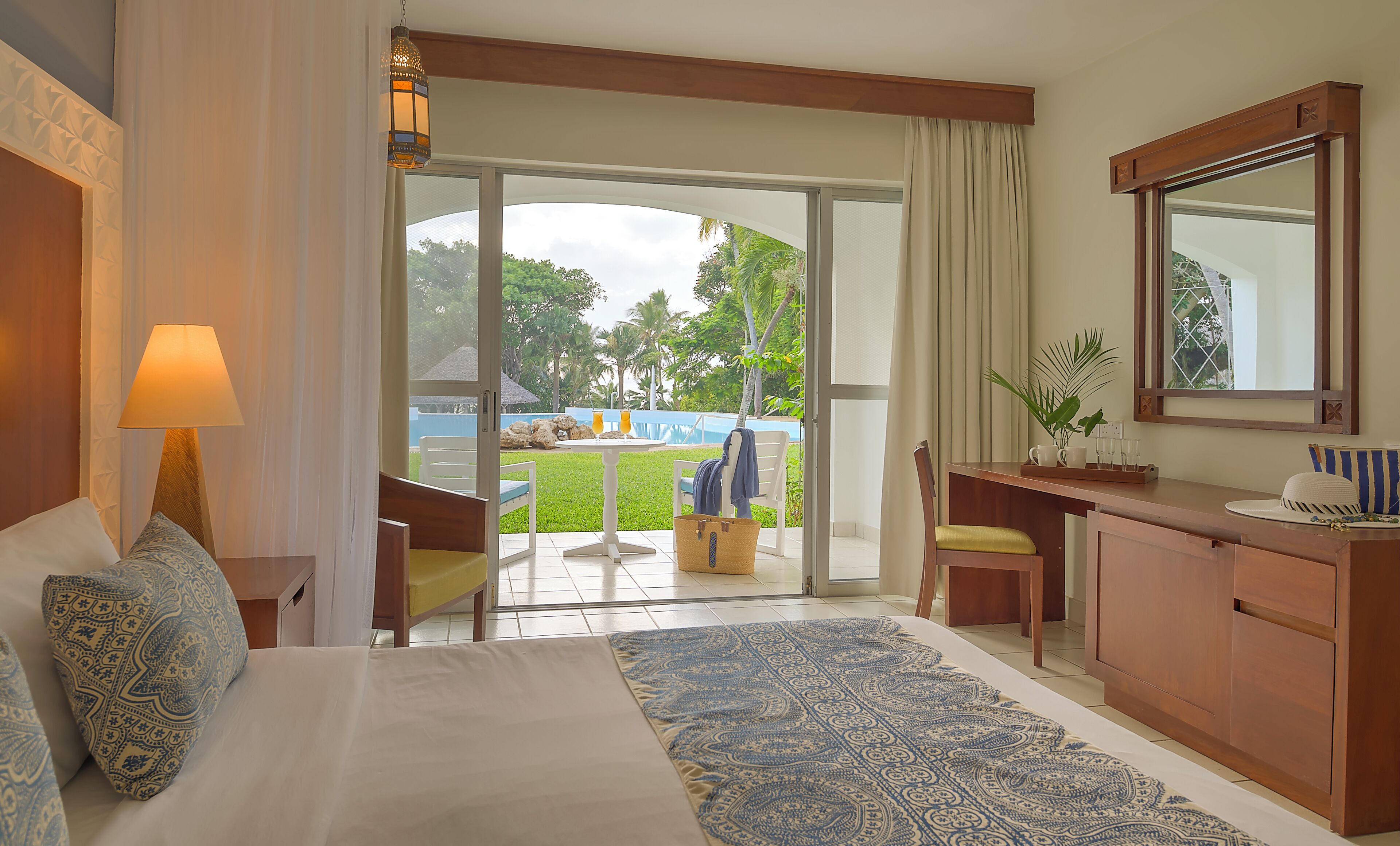 A hotel room with a bed, a desk, a chair, a lamp, and a view of the outdoor area.