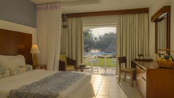 Habitación de hotel con cama, escritorio, silla, mesita y vista a una piscina y zonas verdes al exterior.