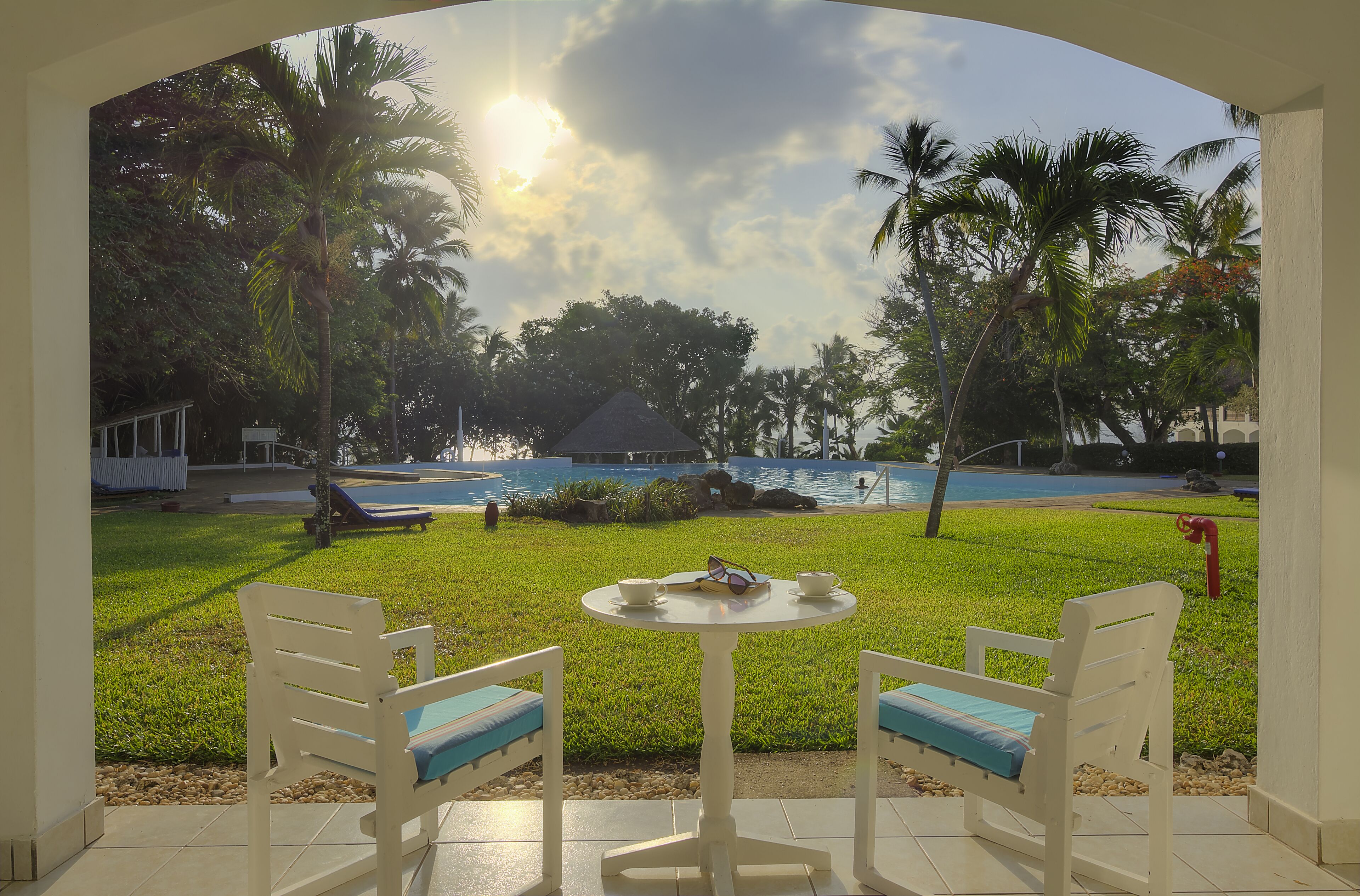 Un patio con dos sillas blancas y una mesita, con vistas a una zona de piscina con palmeras y un cielo despejado.