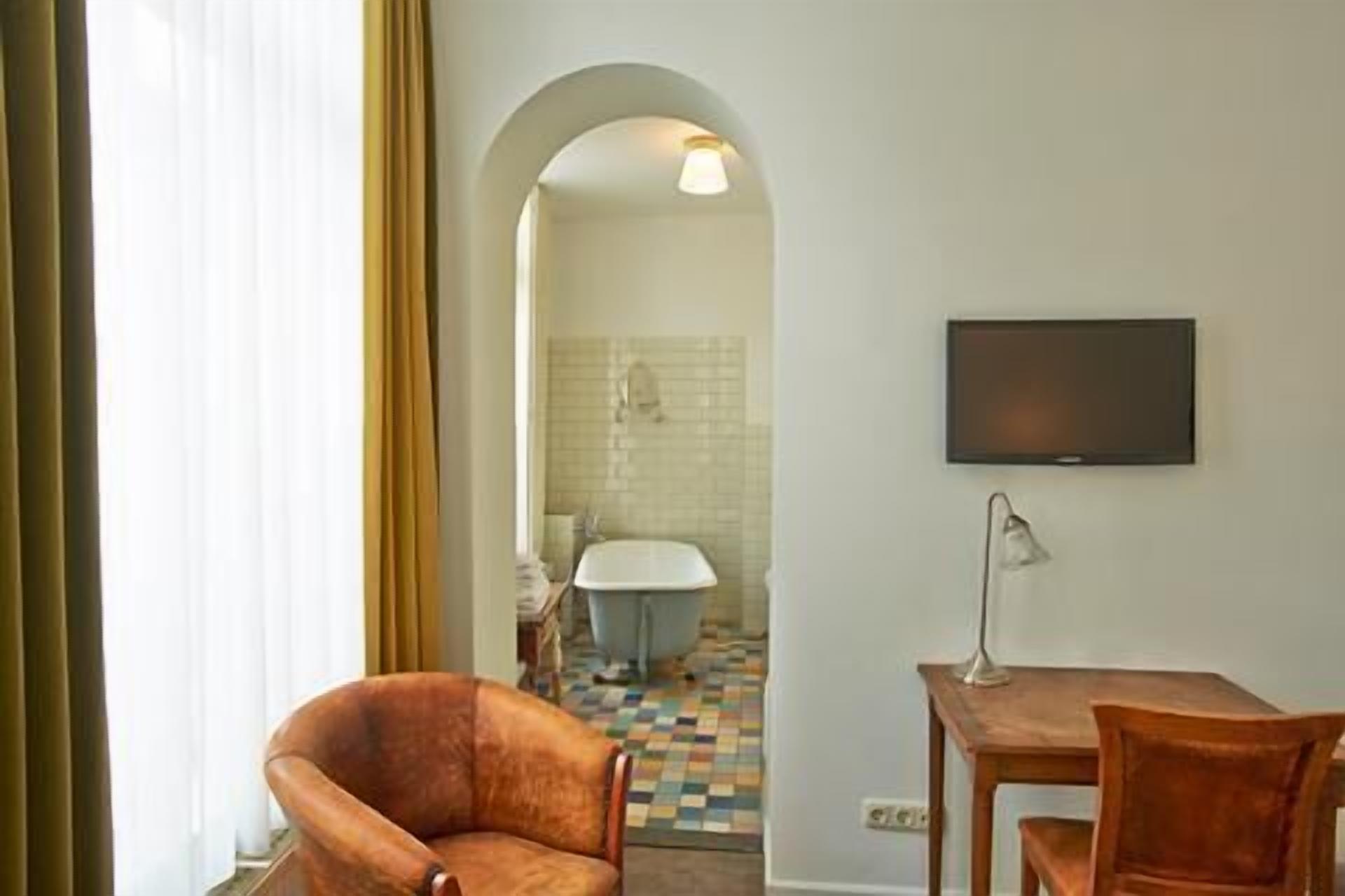 Quarto de hotel com sofá, escrivaninha, televisão e banheiro visível por um arco.