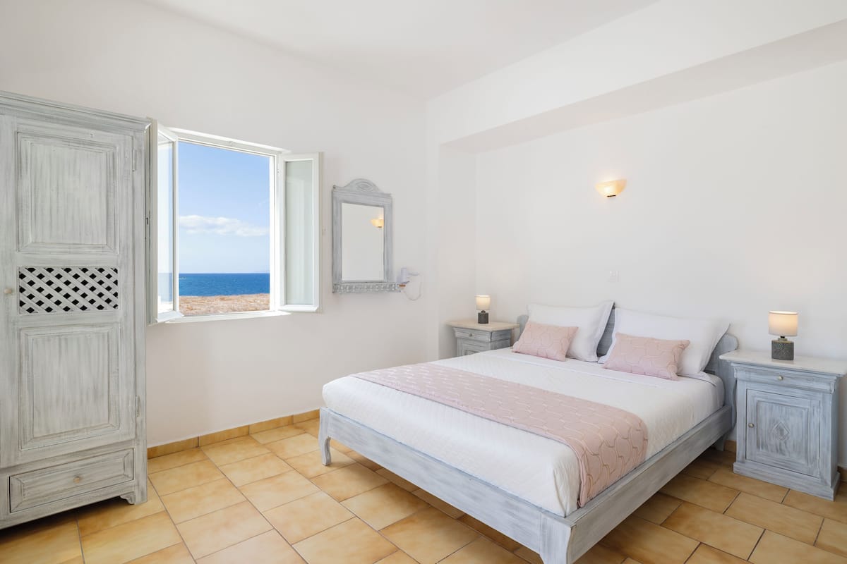 A bedroom with a bed, nightstands, a wardrobe, a window with a view of the sea, and a wall-mounted mirror.