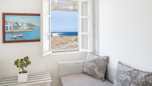 A room with a large window offering a beach view, a framed painting of a coastal scene, and a potted plant on a side table. - Vrachia Studios & Apartments (Santorini)