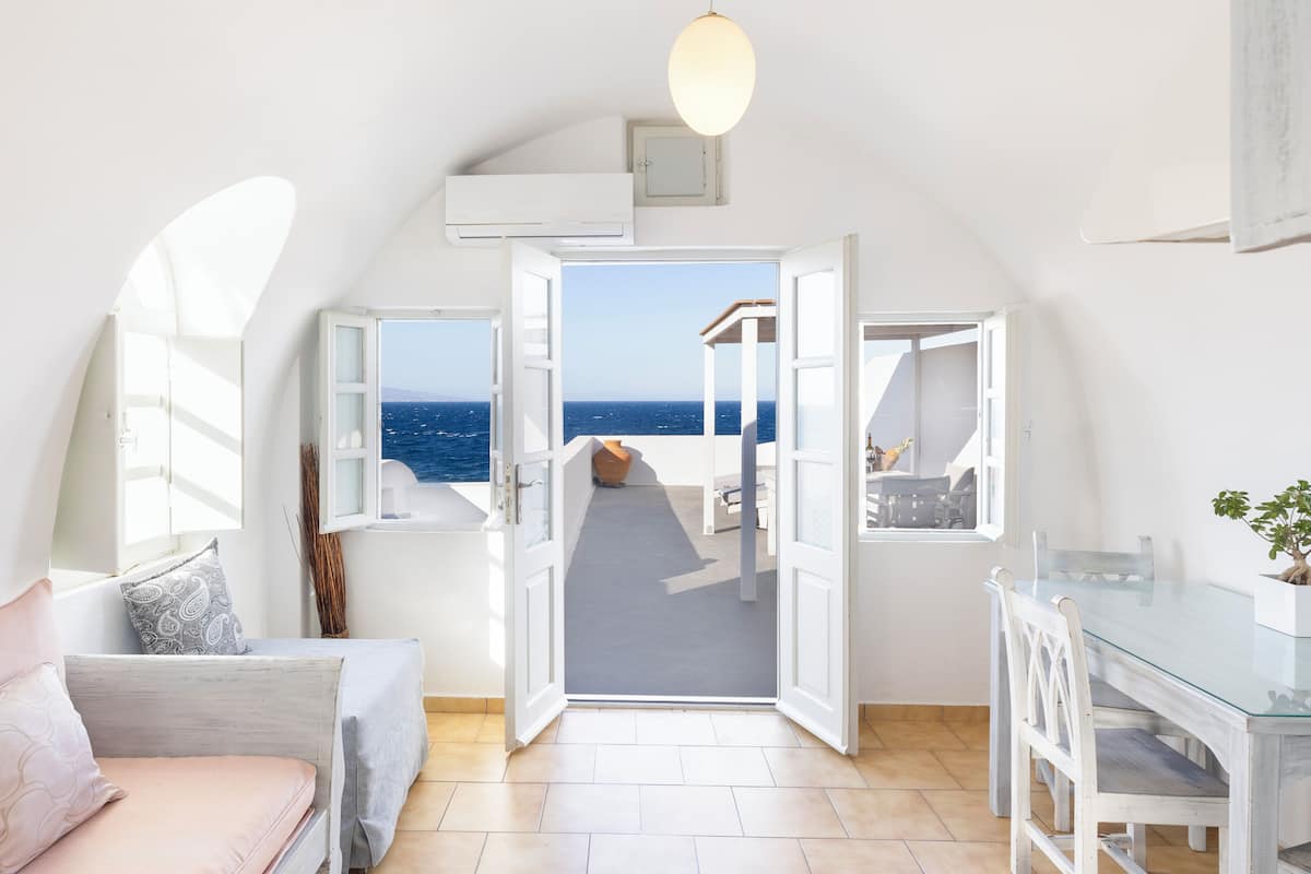 A room with a view of the sea, an open door leading to a balcony, a dining table, and a sofa.