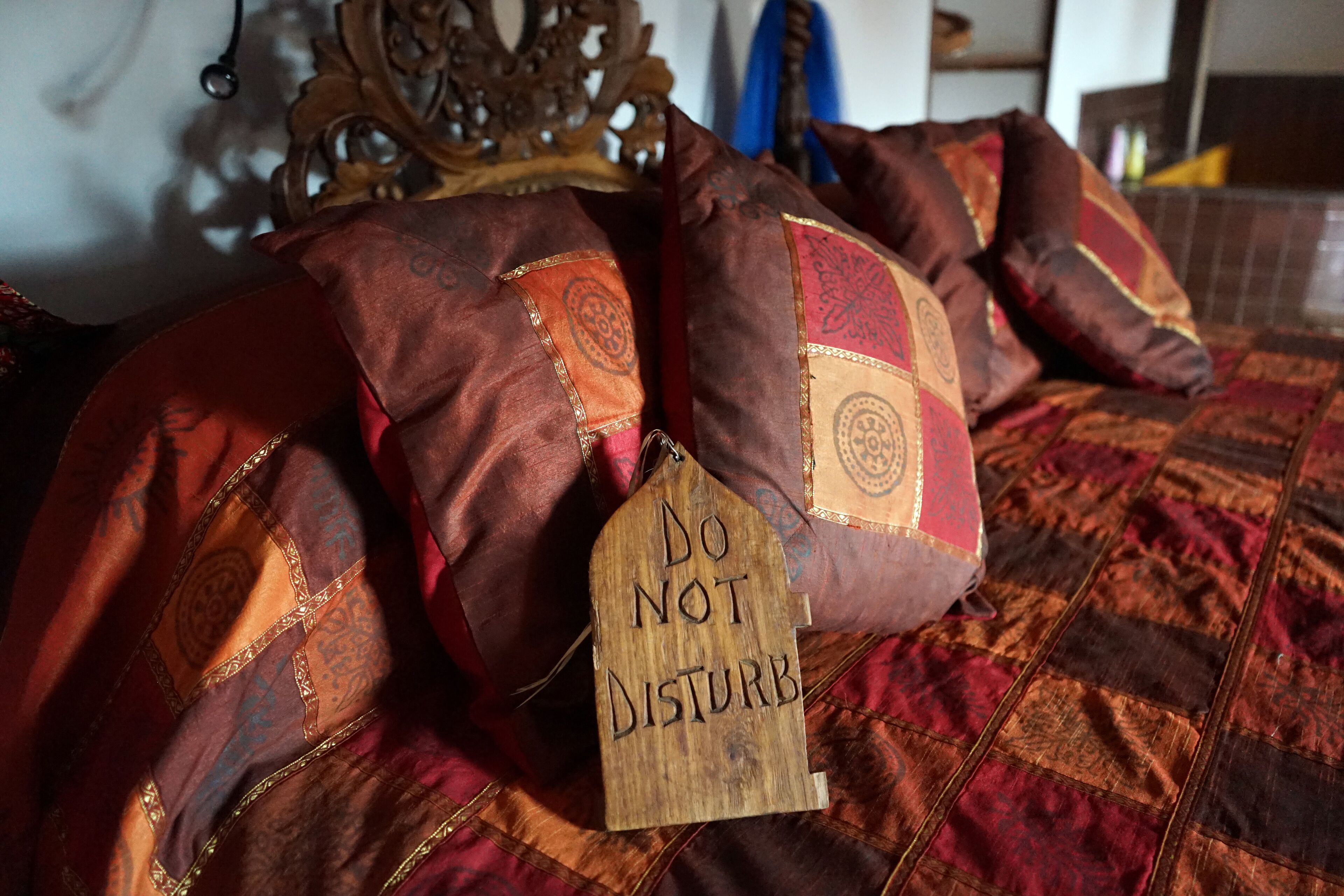 A bed with a red and brown patterned bedspread and pillows with intricate designs. A wooden sign on one pillow reads "DO NOT DISTURB".