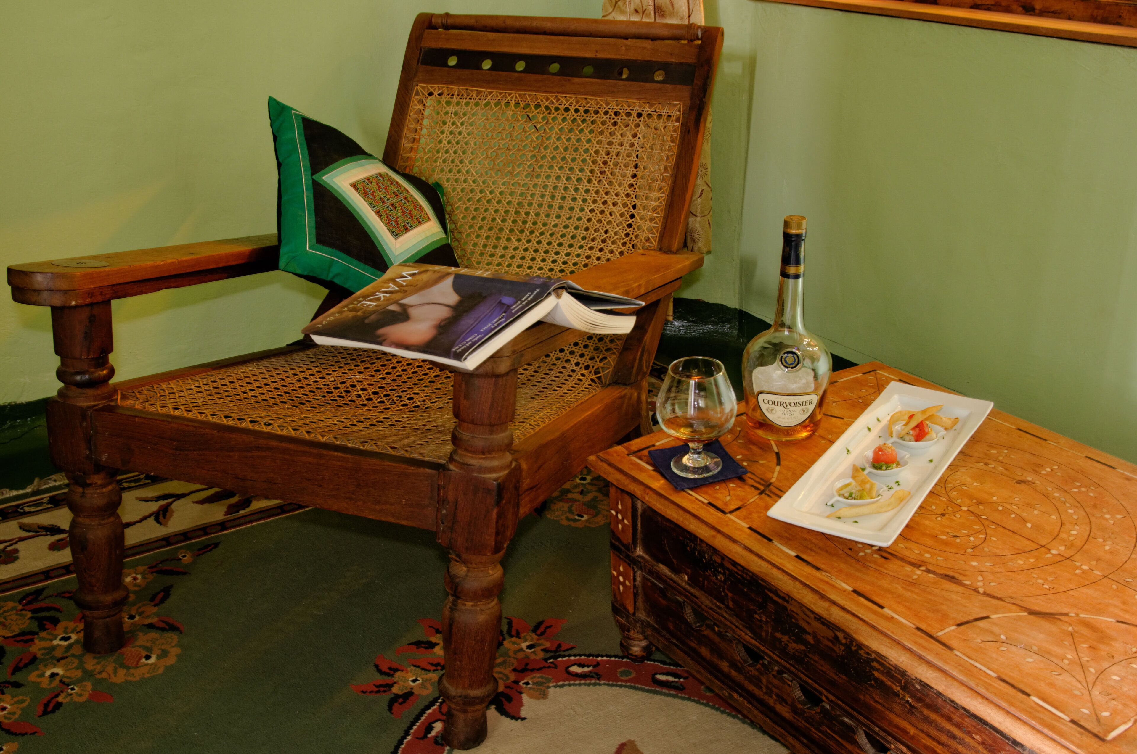Una sedia di legno con schienale intrecciato, un libro, una bottiglia di liquore e un piatto con cibo su un tavolo di legno.