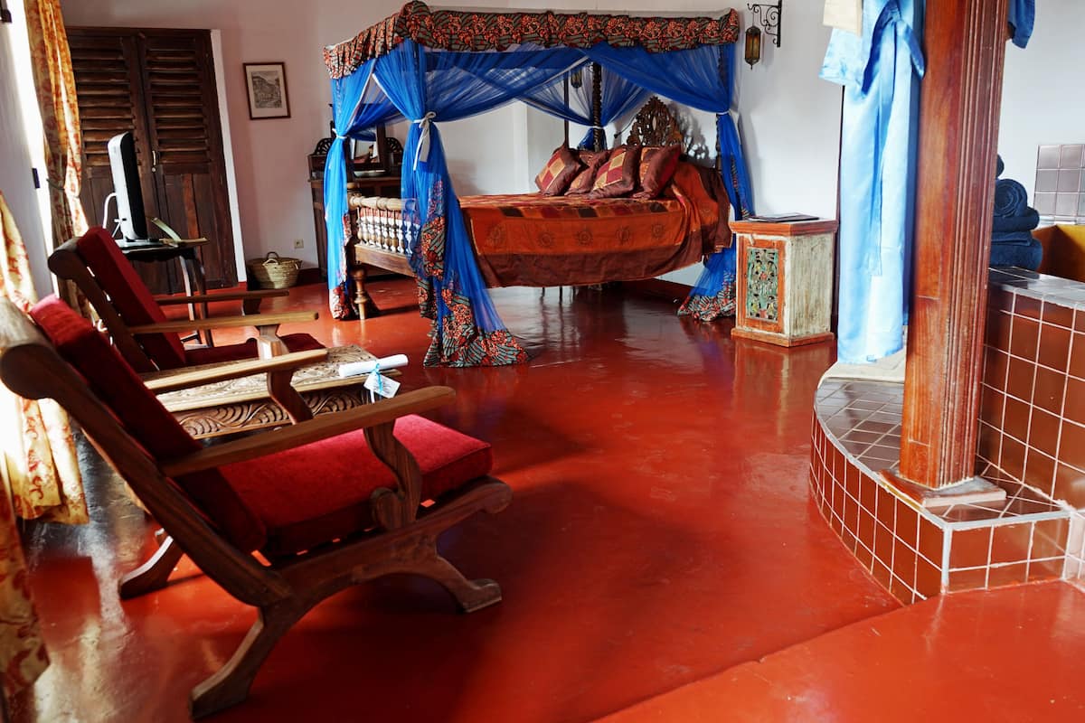 Una habitación con una cama bajo un dosel azul, sillas de madera, una mesita pequeña y suelo de baldosas.