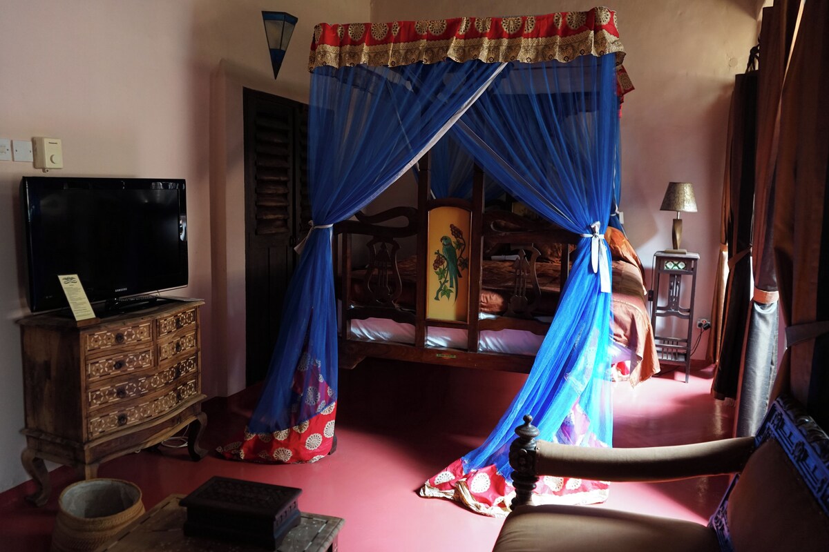 Een slaapkamer met een bed onder een blauw hemeltentje, een houten dressoir, een televisie en een lamp.