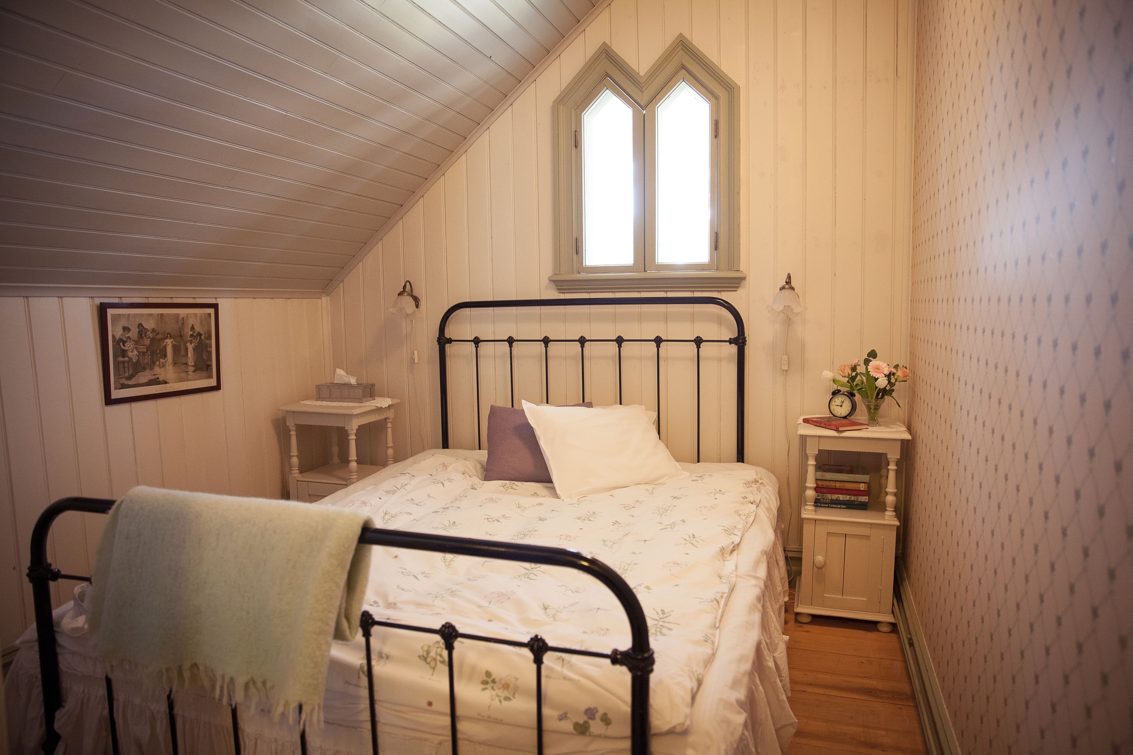 Ein Schlafzimmer mit geneigter Decke, einem Bett mit Metallgestell, einem Fenster mit dekorativem Rahmen, einem Nachttisch mit einer Uhr und Büchern sowie einem Wandbild.